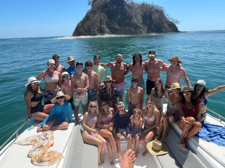 A large private charter group fills the catamaran's bow in front of the rock island off Nosara.