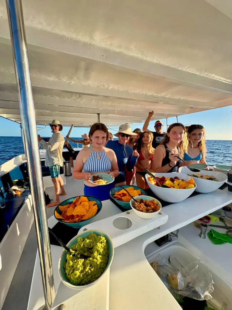 Bowls of guacamole, fruit and salads fill the counter while guests gather on deck behind.