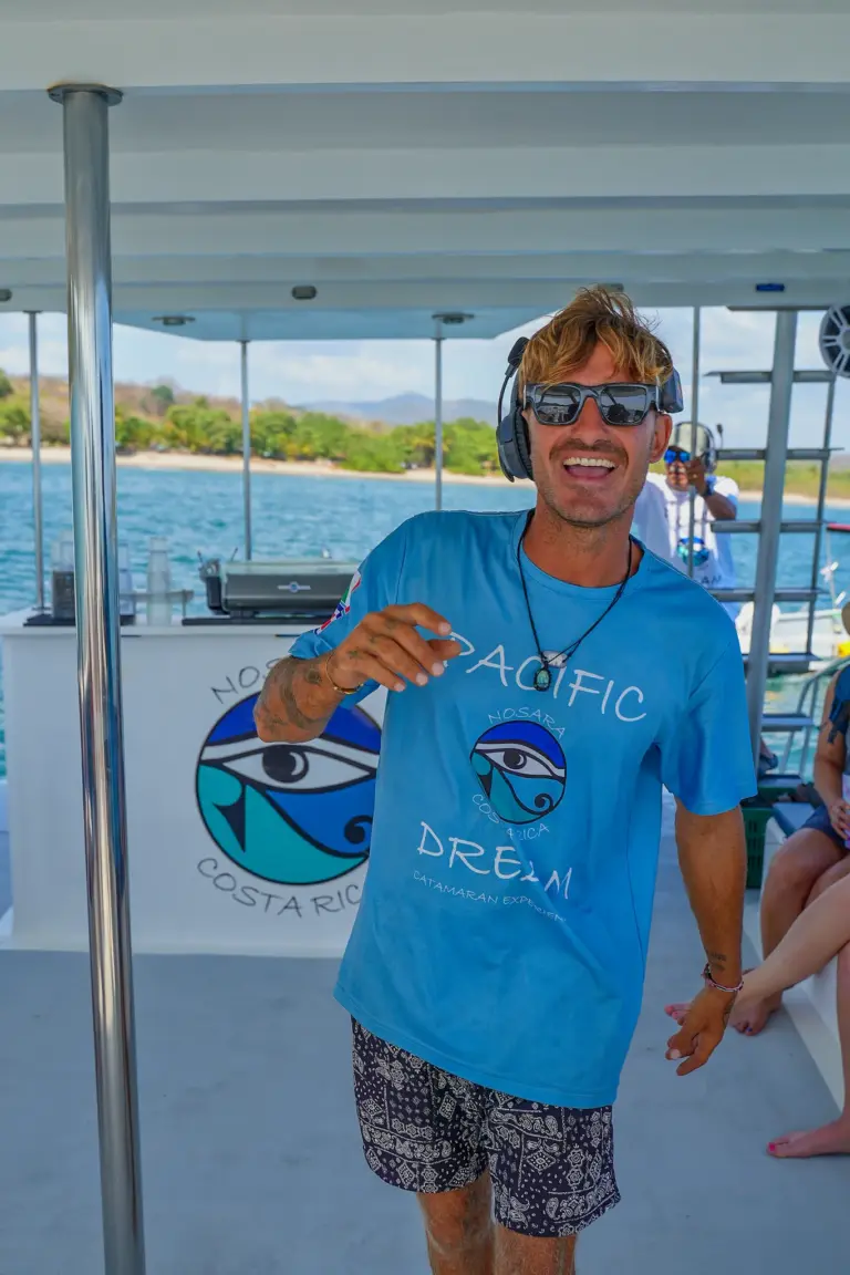 A crew member stands on the catamaran deck with the green Guanacaste coastline behind.