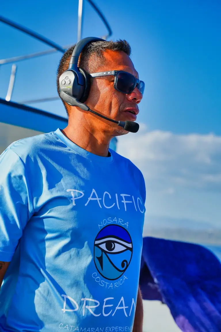 A crew member with a headset microphone talks to guests on the catamaran deck.