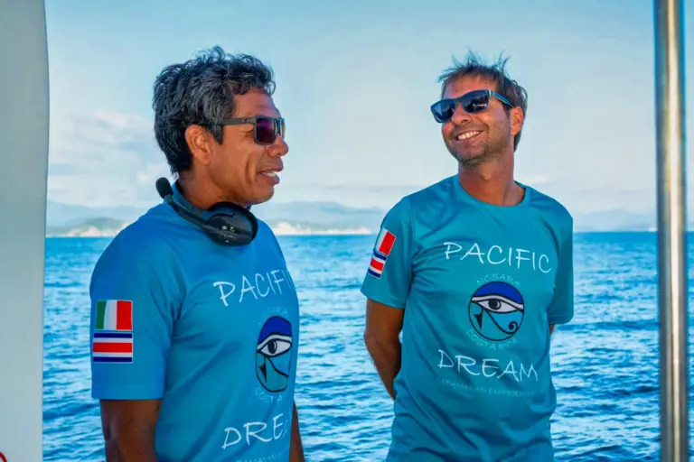 Two crew members stand together on deck with the ocean and coastline behind them.