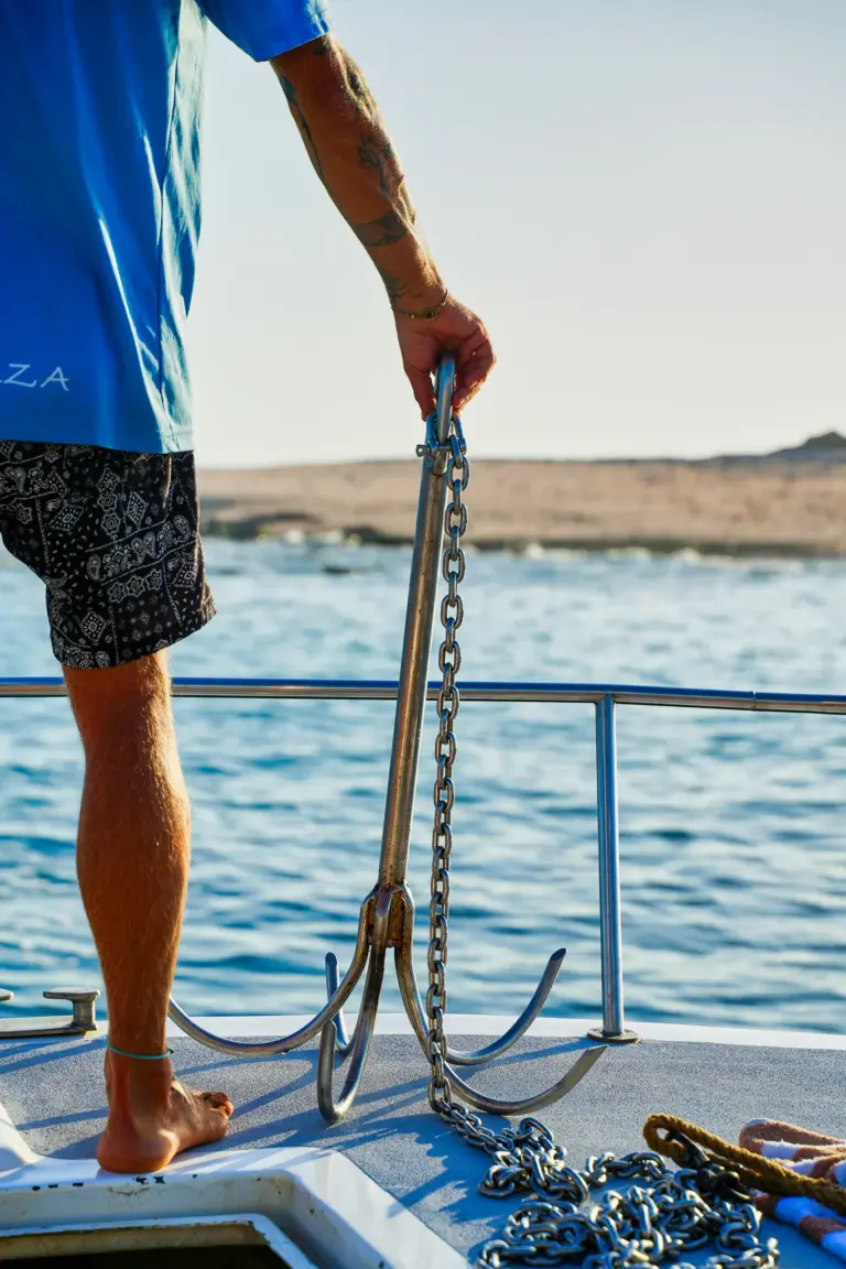 A crew member lowers the anchor chain from the catamaran's bow beside a rocky shore.