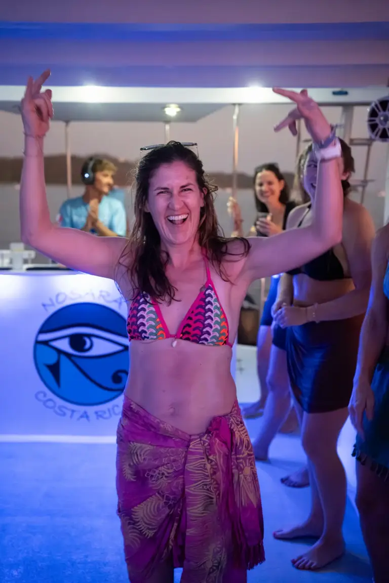 A guest dances with her arms up on the catamaran's lit deck while others join in behind.