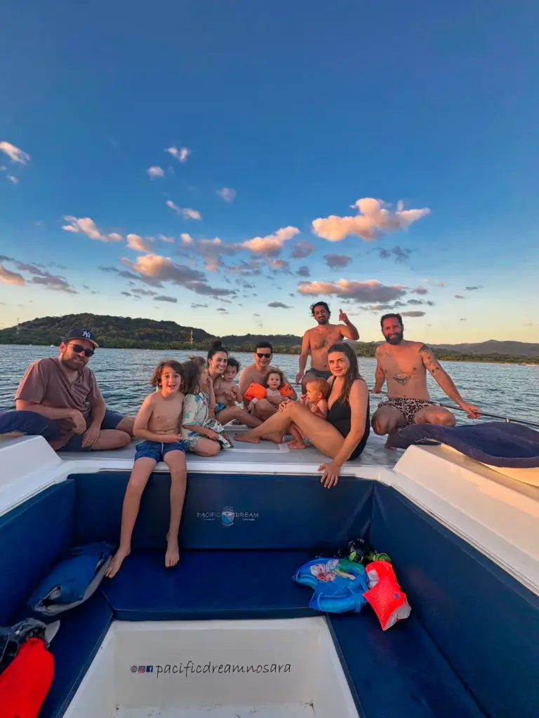 A family group sits along the catamaran's stern with a green headland in the evening light.