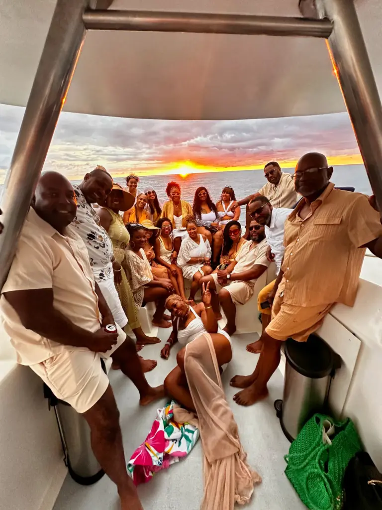 A large group celebrates together on the catamaran's deck as the sun sets behind them.