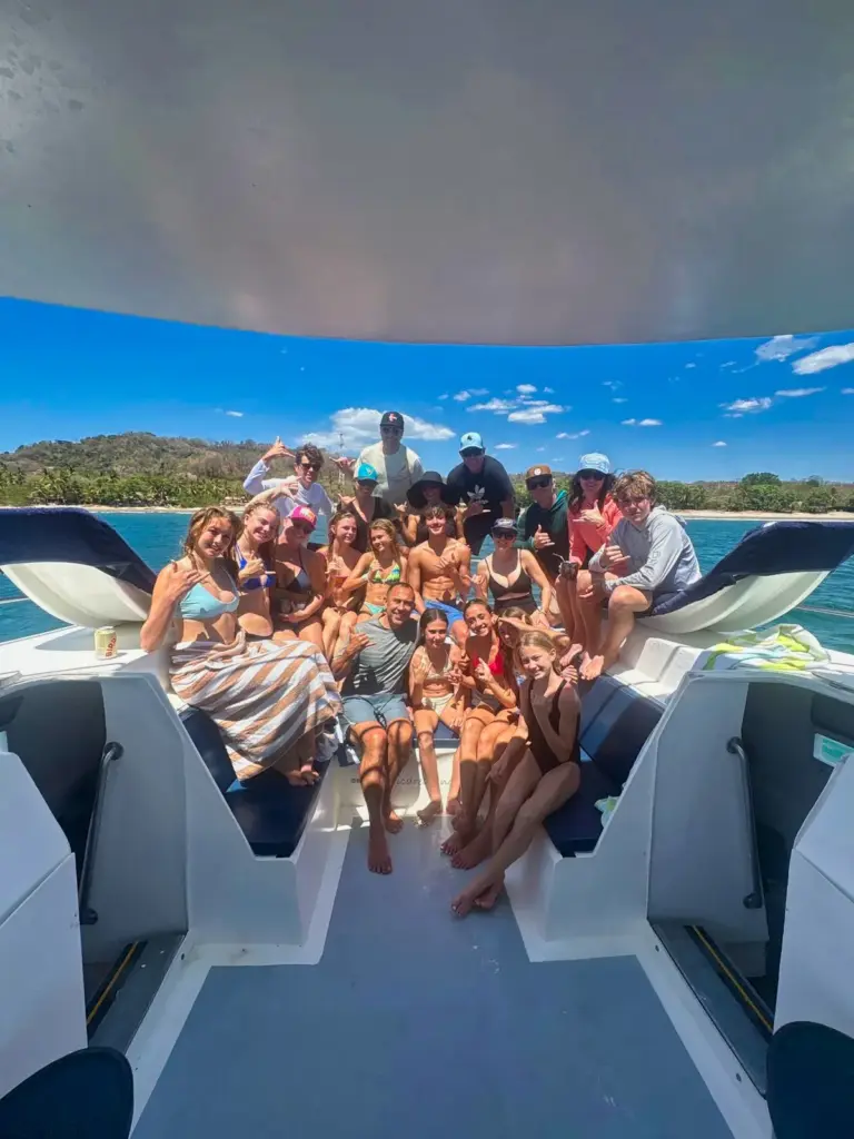 A large group poses together on the catamaran's stern with the green coastline behind.