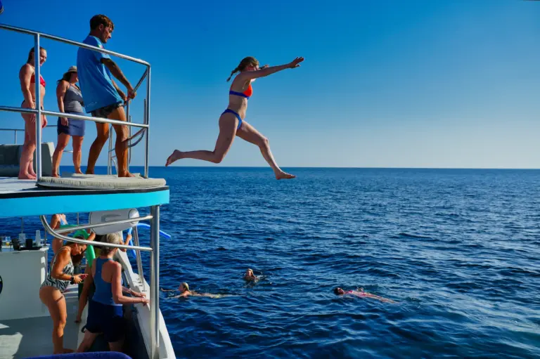 A guest leaps from the catamaran's upper deck into the blue water while others watch and swim.