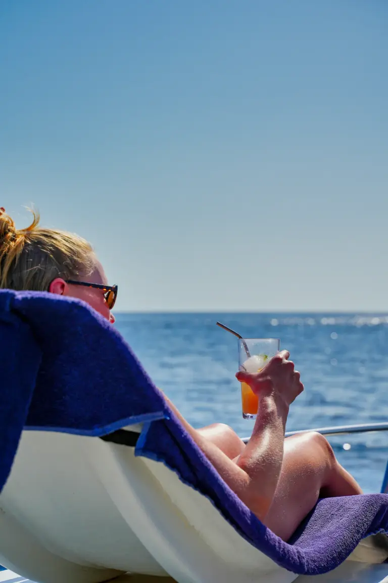 A guest lies back on the catamaran's sun lounger holding a cocktail above the blue water.
