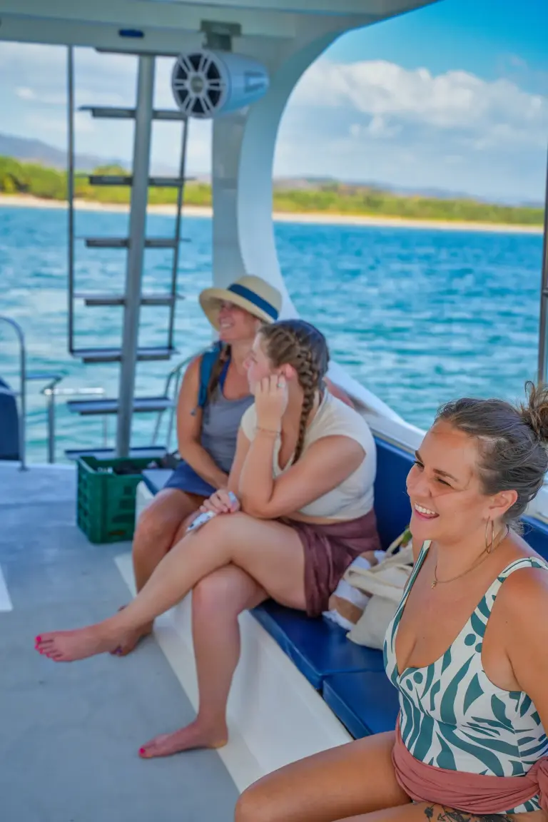 Guests sit and chat on the catamaran's deck with a pale beach and green hills behind.