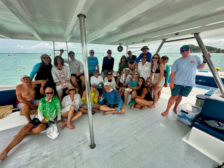 A large group of guests poses together on the catamaran's shaded deck near the coast.