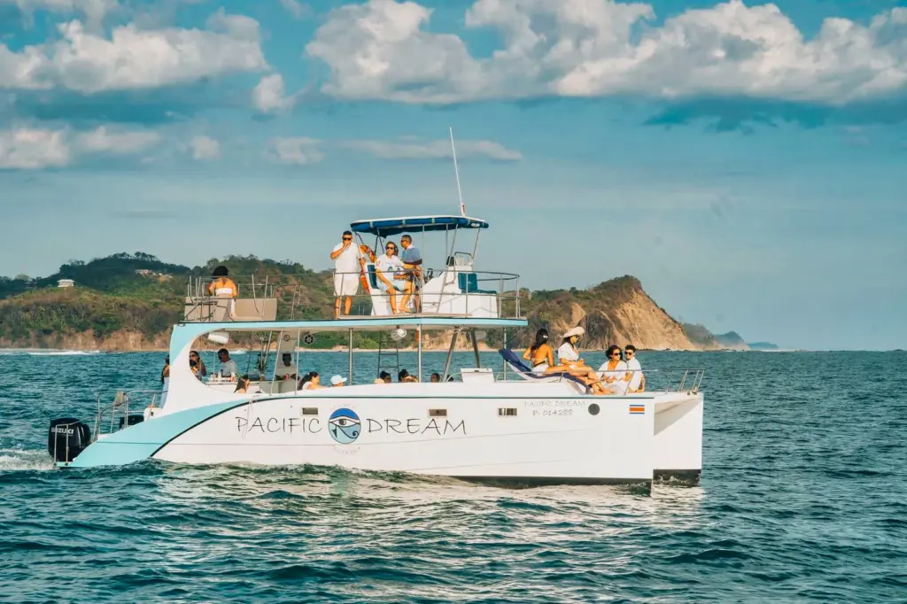 The catamaran cruises past a green headland with guests on the upper deck near Nosara.