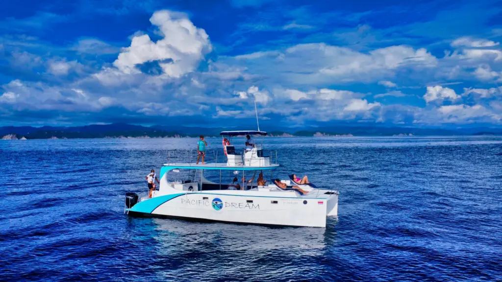 The catamaran cruises with guests on deck under a bright cloud-streaked sky.