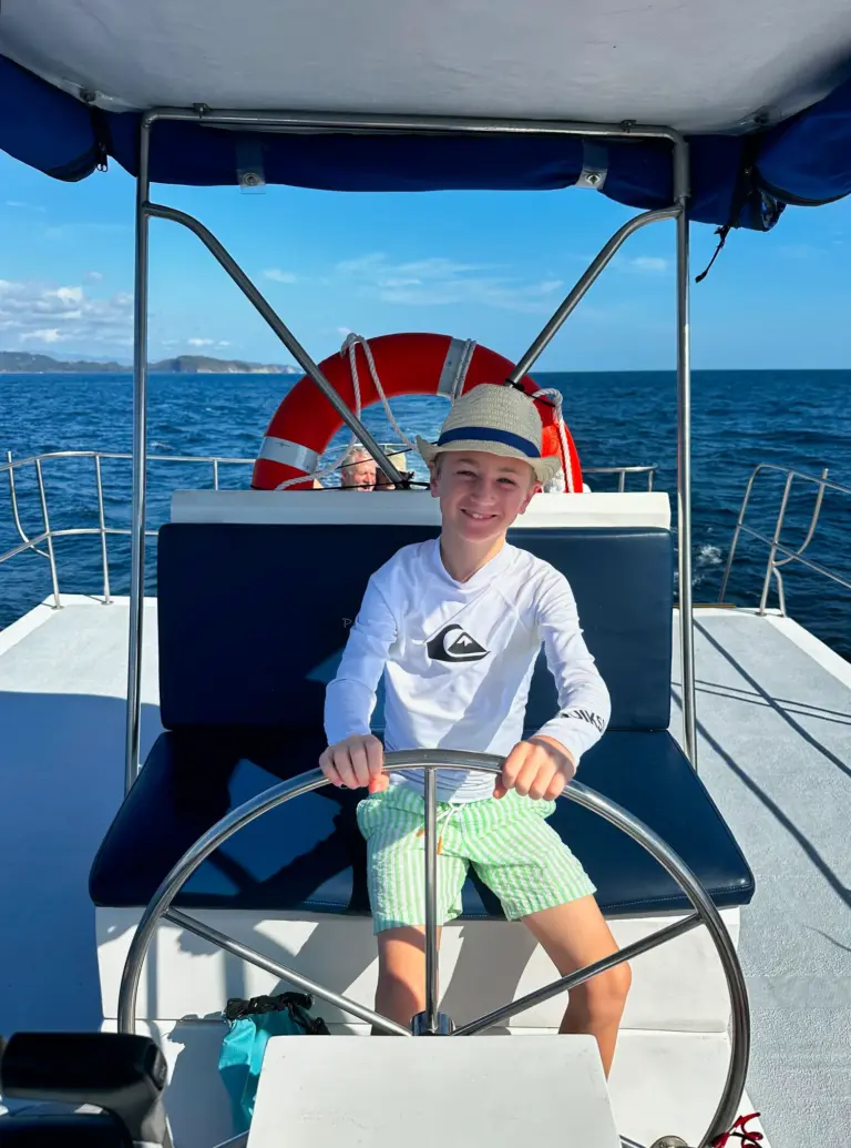A boy in a sun hat takes the catamaran wheel under the shade canopy off Nosara.