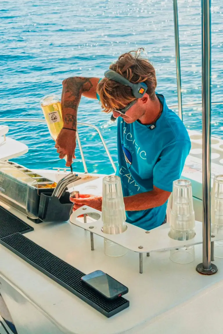 A crew member sets up glasses at the onboard bar with the Pacific Ocean behind.
