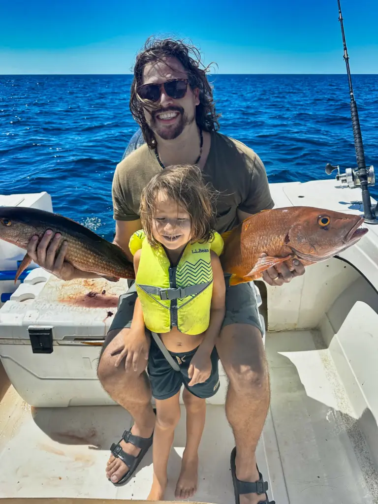 A guest holds two snappers while his daughter in a life jacket smiles on the boat's deck.