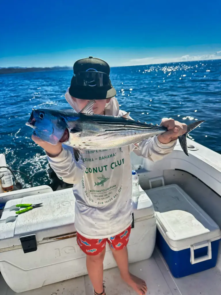 A guest holds up two fish, one in each hand, on the bow with the coastline behind.