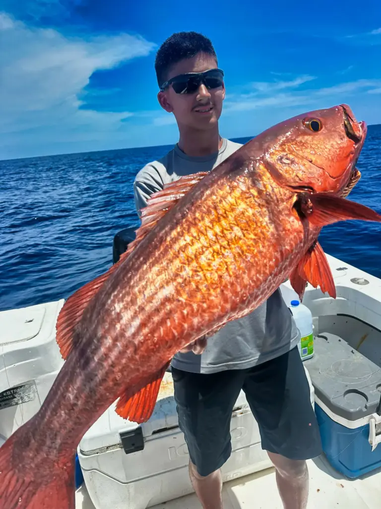 A crew member holds a large snapper across the deck with deep blue water behind.
