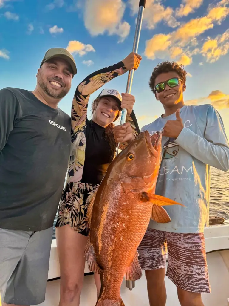 Guests and a crew member hold a snapper on a gaff as evening light hits the water.