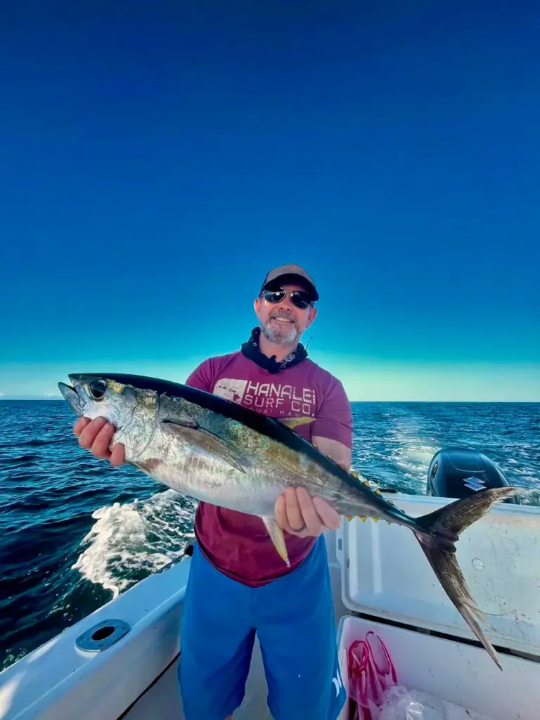 An angler holds a yellowfin tuna at the stern with deep blue Pacific water behind.