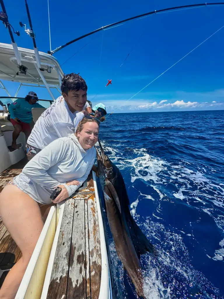 A guest reaches over the transom toward a sailfish held in the water before release.
