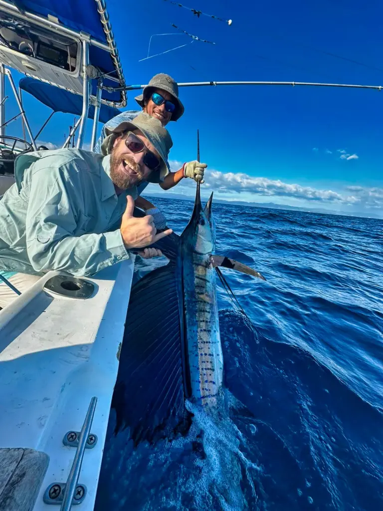 A guest gives a thumbs up next to a sailfish held at the boat's side before release.