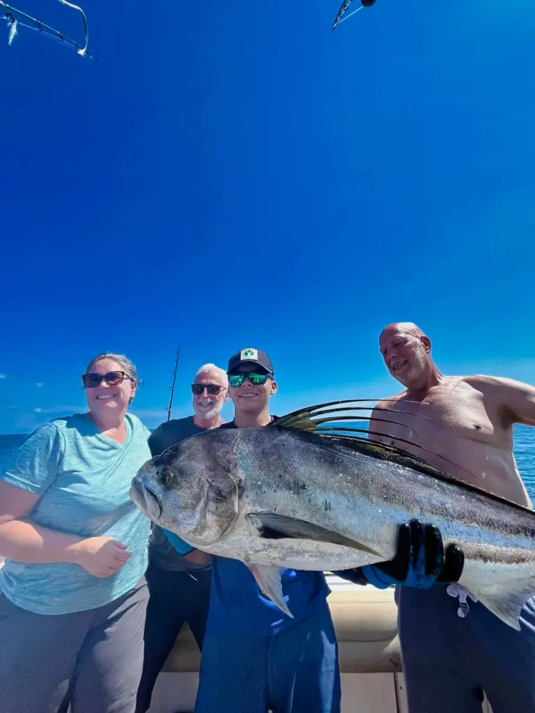 Guests hold a large roosterfish along the rail with deep blue water behind the boat.