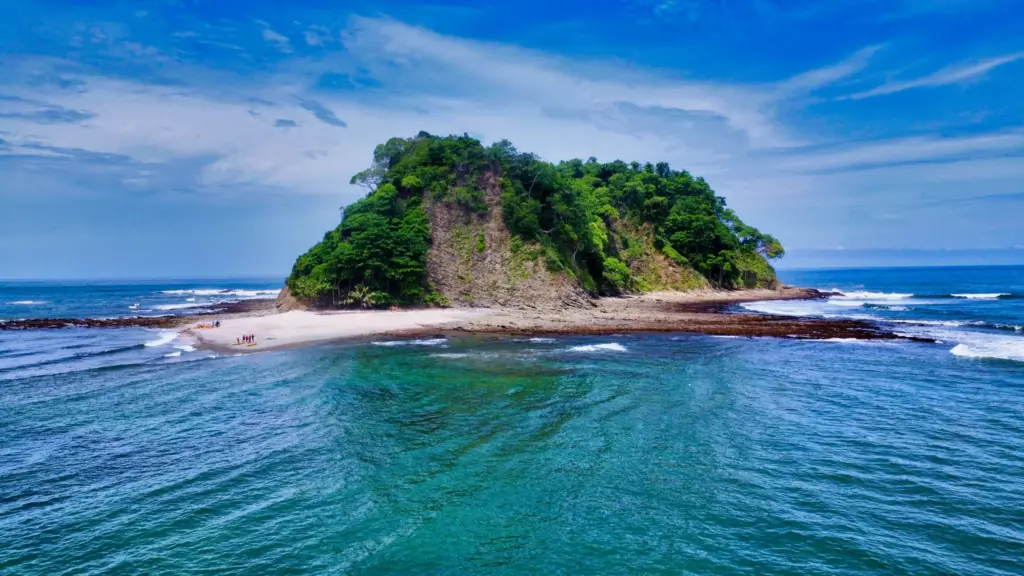 Aerial view of a green island joined to a curved sand spit in clear water near Nosara.