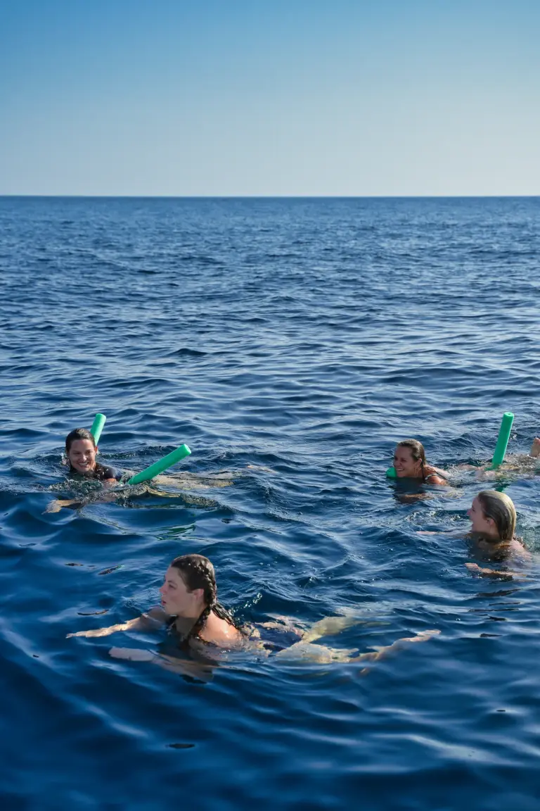 Four guests swim and float with pool noodles in deep blue water away from the boat.