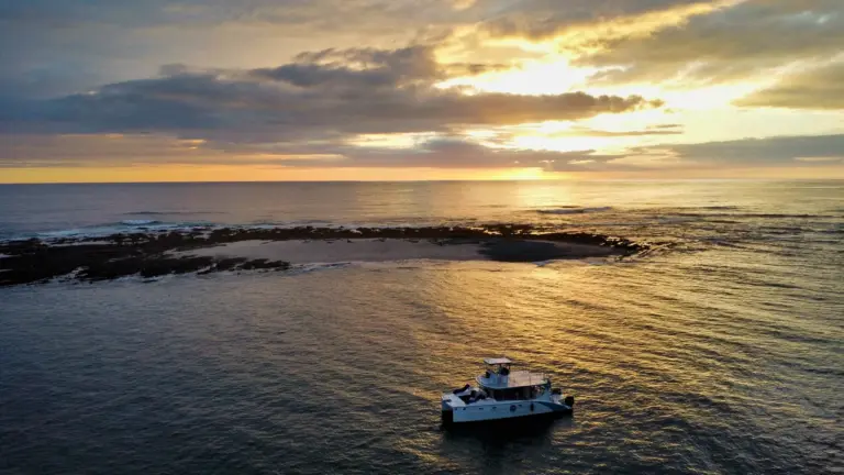 Drone view of the catamaran anchored beside a reef as the sun drops over the Pacific.