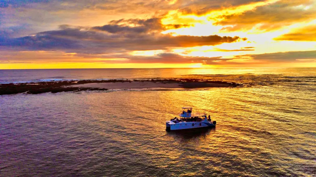 Aerial view of the boat anchored beside a reef under a wide gold and purple sunset.