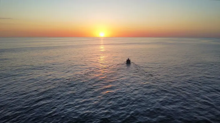 A small boat crosses the calm Pacific under a wide orange sunset seen from the deck.