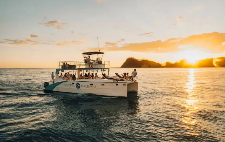 The catamaran cruises past a headland with guests on both decks as the sun sets.