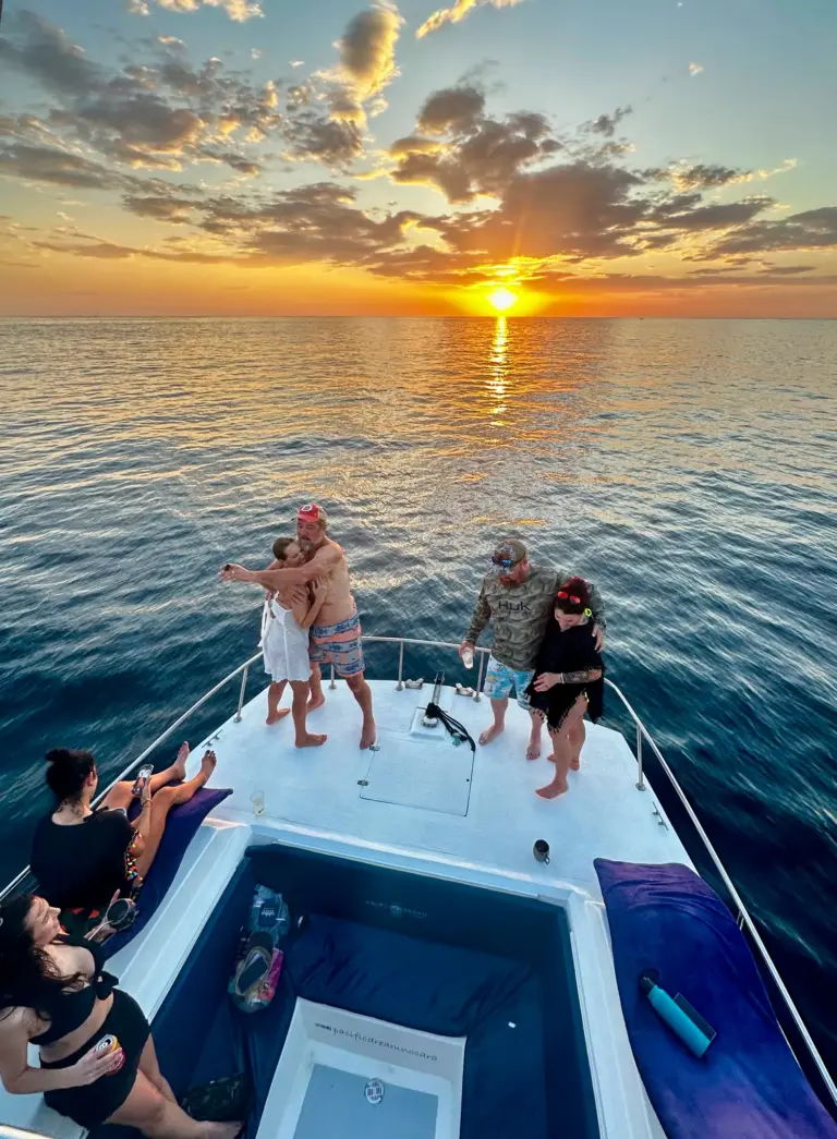A couple hug on the catamaran's bow while other guests watch the sun set over the Pacific.