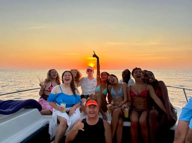 A group of friends laughs and celebrates on the catamaran deck against a bright orange sky.