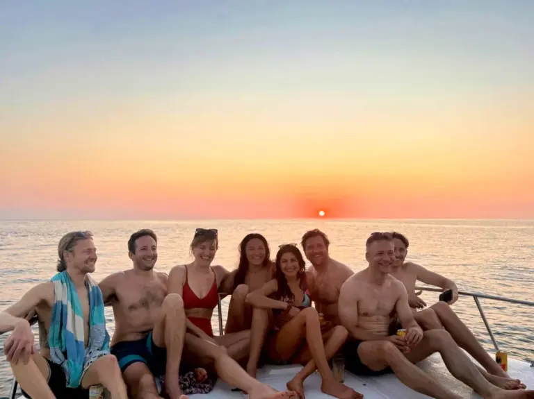 A row of guests sits along the catamaran's side deck against a soft orange sunset sky.