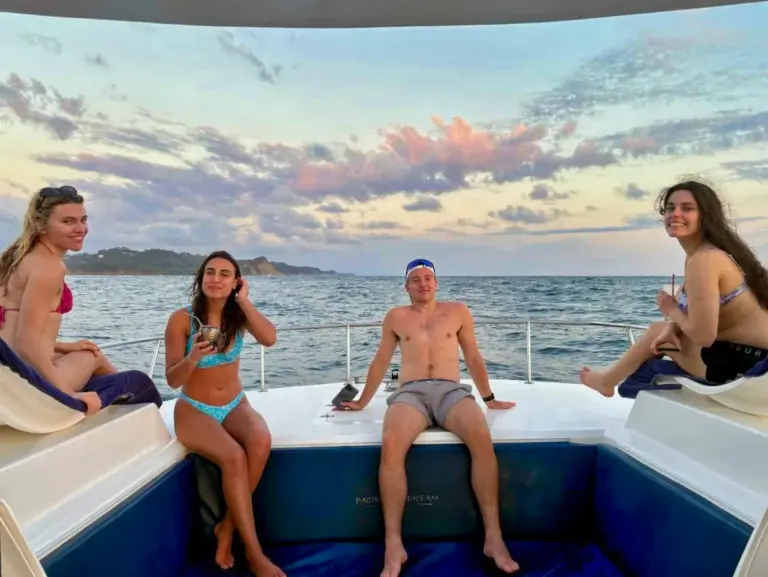 Guests sit and lie on the catamaran's bow cushions under a soft pastel evening sky.