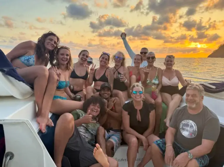 Guests laugh and raise their arms on deck while the sky turns orange behind them.