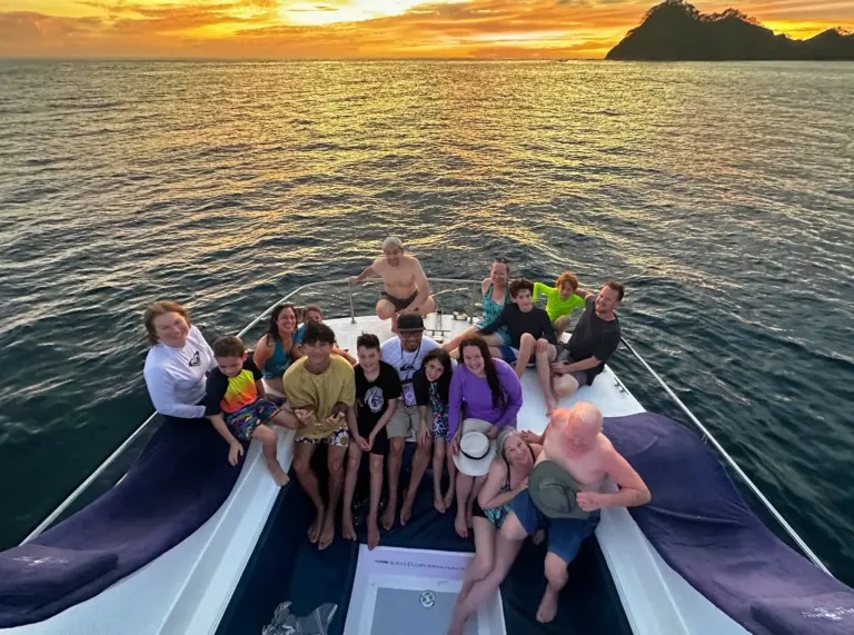 A large group of guests fills the catamaran's bow as golden light spreads across the bay.