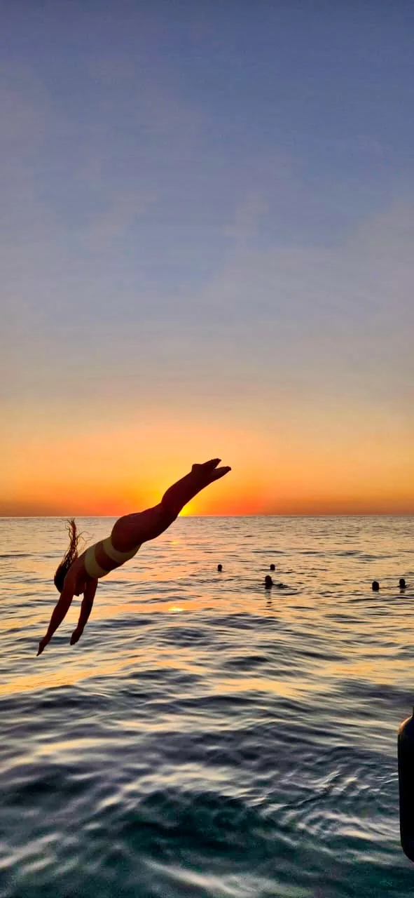 A guest dives from the boat into calm golden water as the sun sets off the Nosara coast.