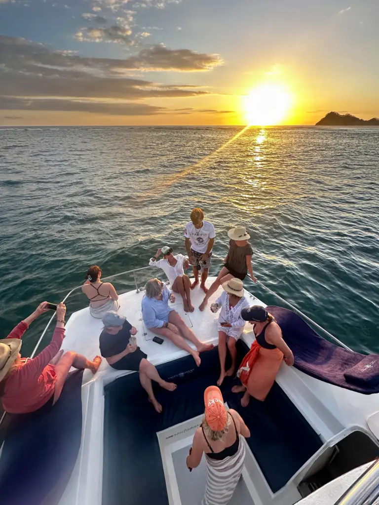Guests spread across the catamaran's bow watching the sun drop behind a headland off Nosara.