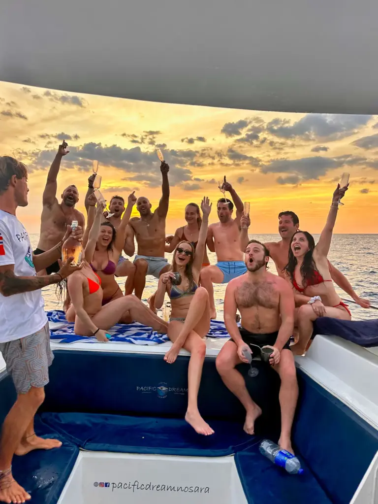 Guests raise drinks and cheer on the catamaran's bow under a dramatic golden sky.