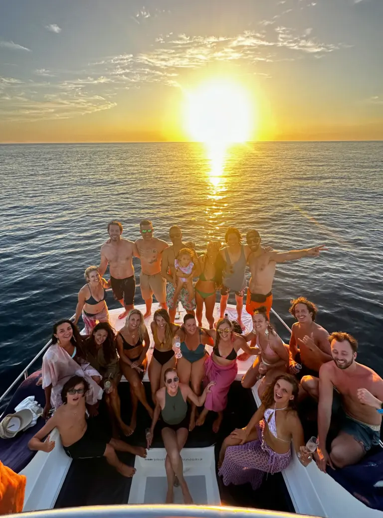 A large group of guests fills the catamaran's bow as the sun burns low over the water.