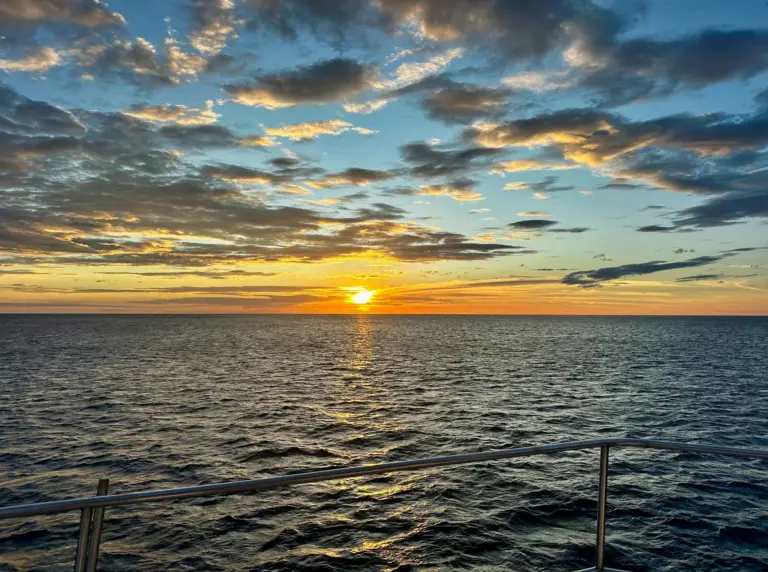 The sun sinks toward the Pacific horizon seen from the boat's rail on a sunset cruise.