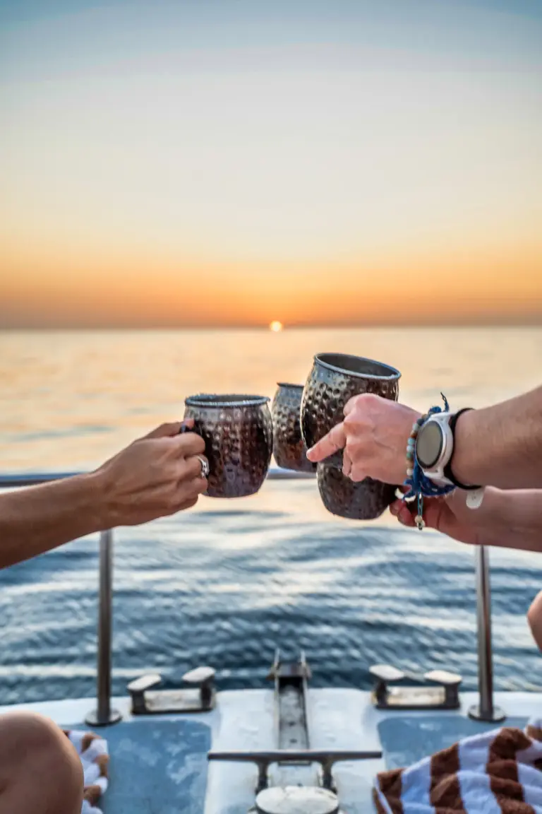 Guests clink copper mugs together at the rail as the sun sets over the Pacific.