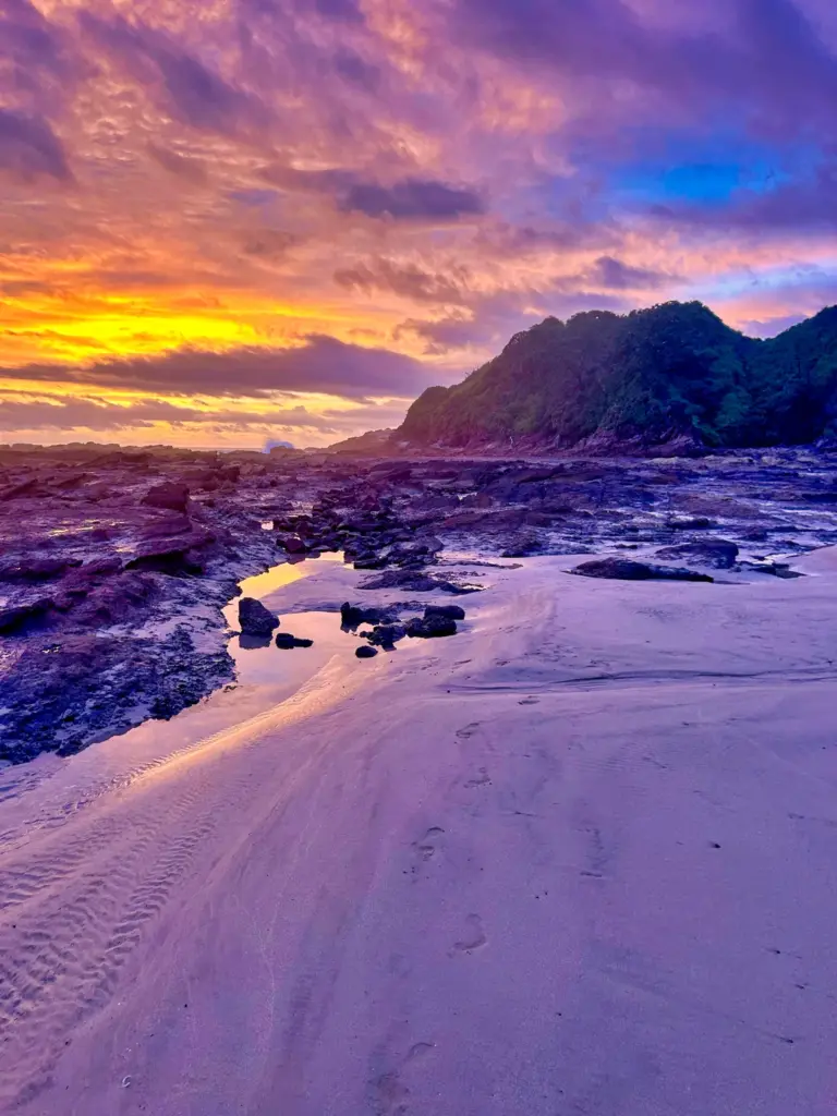 A purple and gold sunset lights the sand and rock ledges of a beach on the Nosara coast.